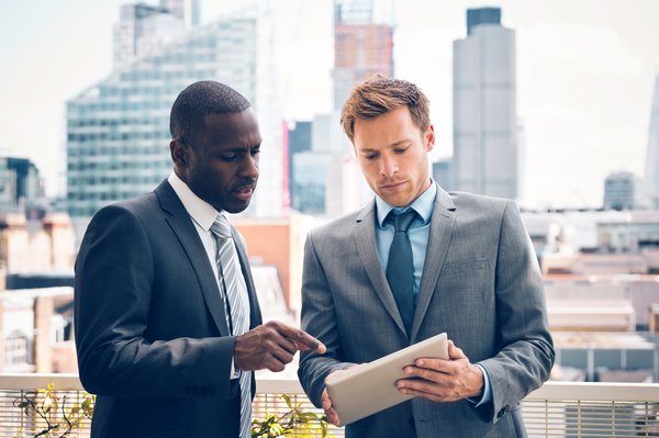 Businesspeople discussing a project on a digital tablet while standing on a balcony with a cityscape in the background.