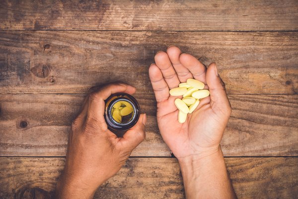 Hands lying on a wooden table and holding some pills in one hand and an open pill bottle in the other.